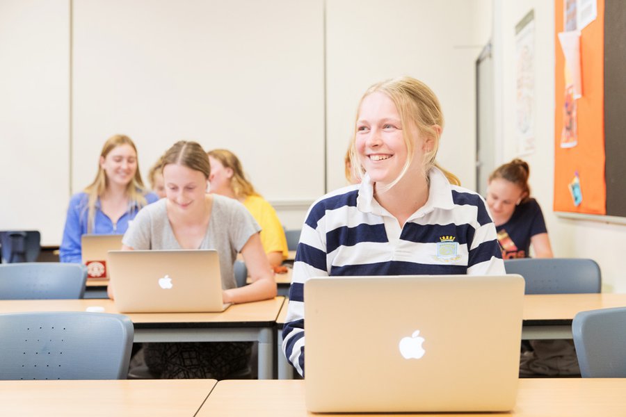 Student in Boarder's Jersey Doing Work on Her Laptop in a Classroom with Other Students Behind Her